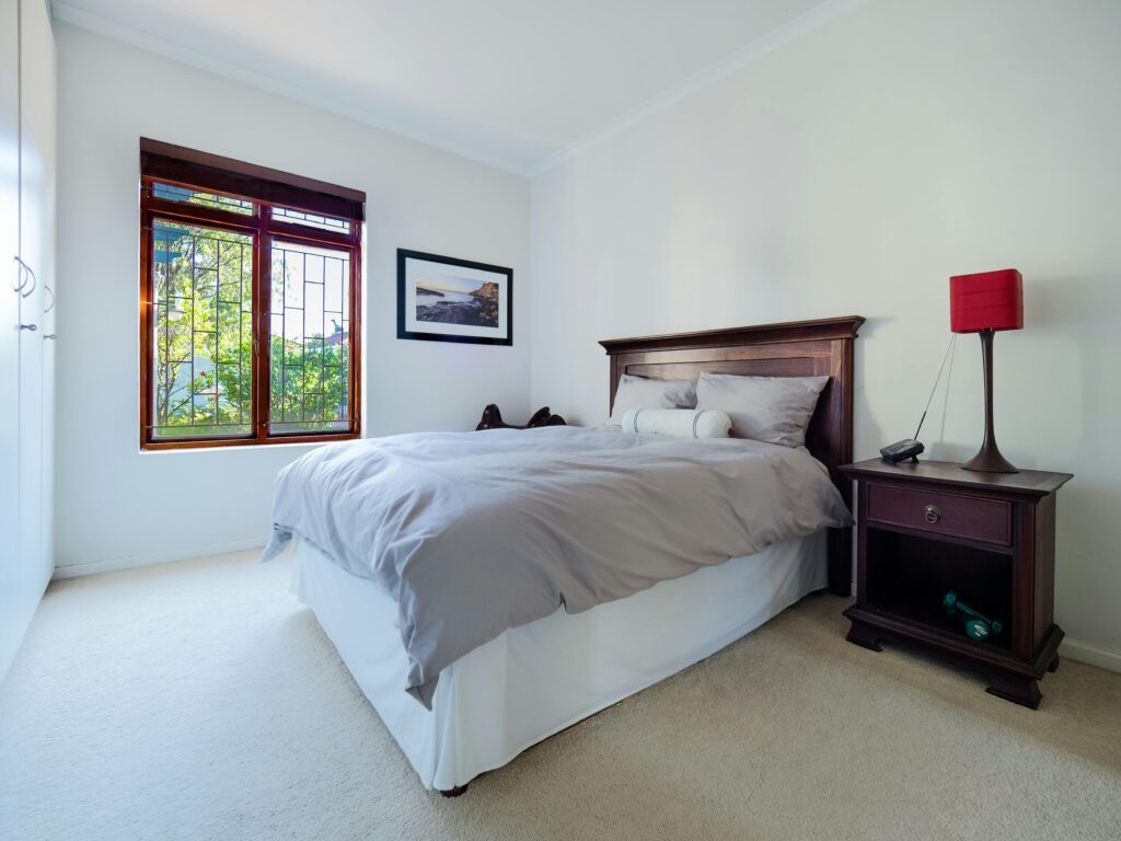 A stylish and comfortable bedroom interior featuring a wooden bed and nightstand with a red lamp and large window.