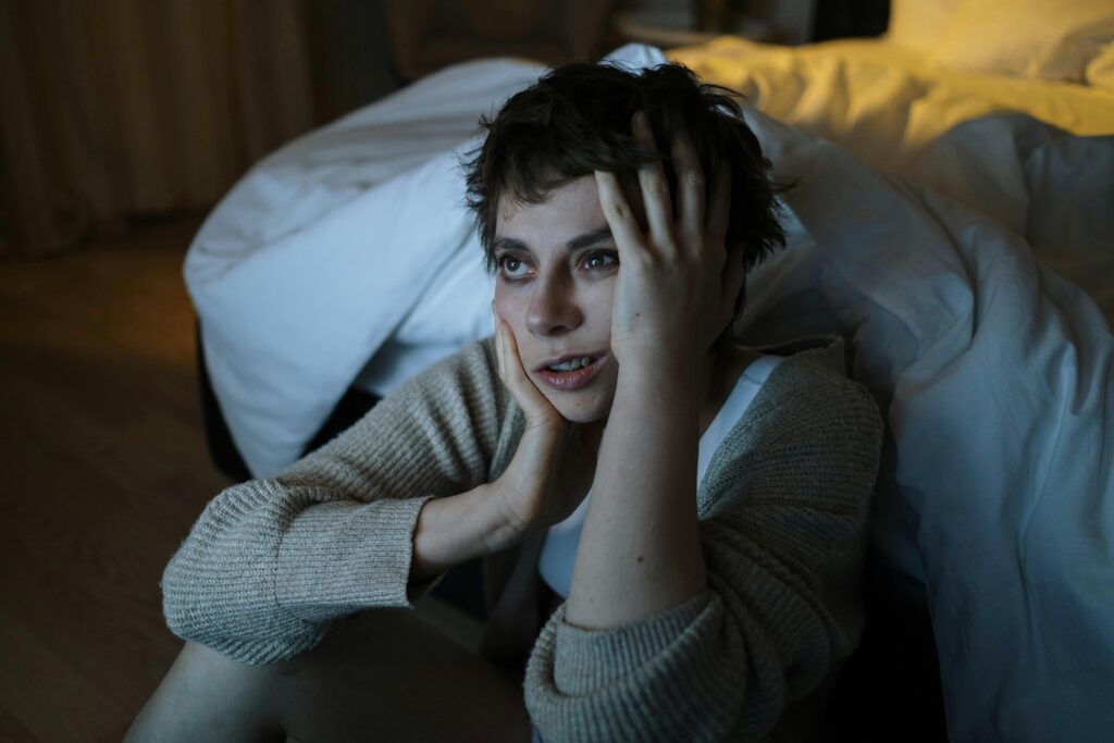 A woman sitting on the floor by a bed with a worried expression, touching her face.